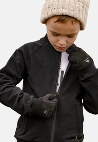 Czarna kurtka na zamek z prążkowanymi mankietami i kołnierzem, noszona na białej koszulce. Czarne rękawiczki z logo, a na głowie teksturowana, beżowa czapka z dzianiny.