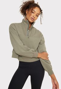 Sudadera corta de color verde oliva con cuello de media cremallera, mangas anchas y puños acanalados, combinada con leggings negros. Textura suave.