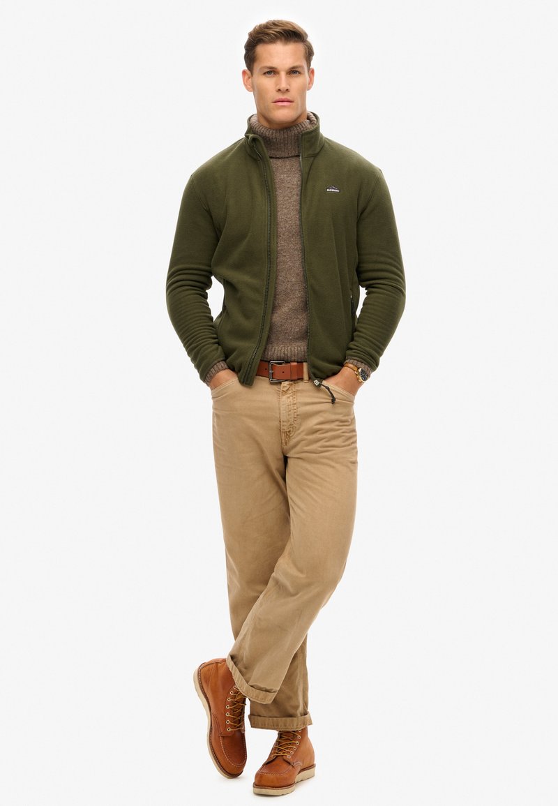 Veste en polaire zippée vert olive sur un col roulé marron, associée à un pantalon beige et des bottes beige. Comprend une ceinture marron et une montre.