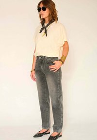 T-shirt beige clair oversize, jean gris taille haute, écharpe noire, ballerines noires avec détails œillets, et bracelets dorés.