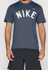 Tummansininen Nike-paita, jossa on lyhyet hihat, ja jossa on suuri valkoinen teksti "NIKE" kaarevassa, kuluneessa fontissa. Valmistettu pehmeästä ja hengittävästä kankaasta.