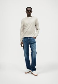 Top beige a maniche lunghe, jeans blu e sneakers bianche. Il modello indossa occhiali da sole scuri. Design semplice con una vestibilità rilassata e colori classici.