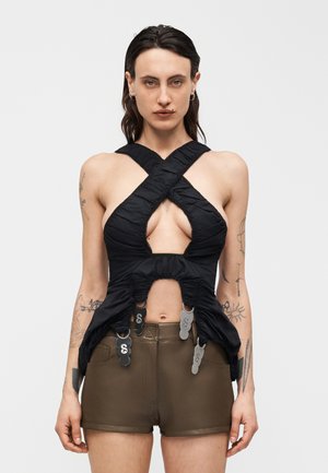 Γυναίκα με τατουάζ που φοράει μαύρο τοπ με λουριά σε σχήμα Χ και μεταλλικά κλιπ, καθώς και καφέ δερμάτινα σορτς, στέκεται μπροστά από απλό φόντο.