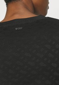 Pull noir avec un motif texturé, un col côtelé et un petit logo argenté à l'arrière près du col.