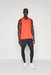 Homem com uma t-shirt atlética sem mangas laranja, calções pretos sobre leggings pretos, ténis brancos e grandes óculos de sol desportivos, de pé contra um fundo simples.