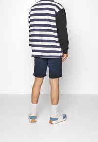 Bakifrån vy av en person som bär en randig långärmad skjorta i marinblått och vitt, med svarta ärmar, matchad med mörka denimshorts och sneakers.