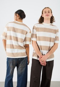Striped short-sleeve T-shirt featuring alternating beige and white stripes, with a crew neck and relaxed fit, worn with blue and brown pants.