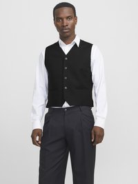 Black tailored waistcoat with five buttons and pockets, worn over a white dress shirt, paired with dark trousers.