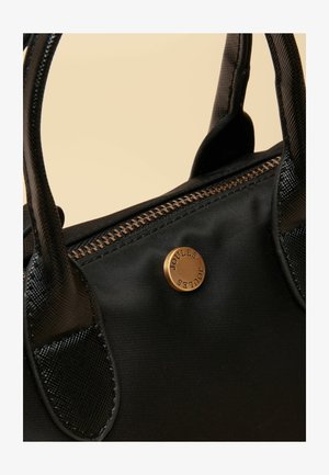 Zwarte nylon handtas met twee korte handvatten, ritssluiting en een rond messing logobadge. Heeft een gladde textuur en contrasterende stiksels.