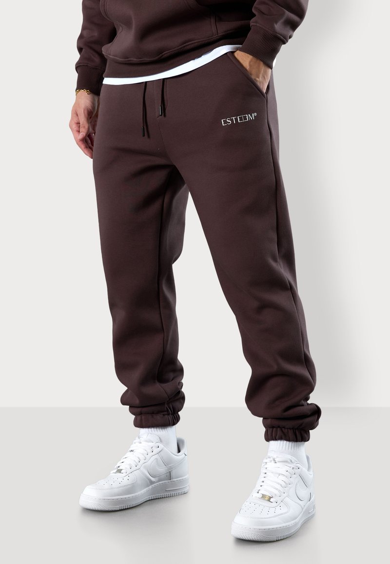 Braune Jogginghosen mit elastischen Bündchen, Kordelzugbund, seitlichen Taschen und weißem "ESTEEM"-Logo. Tragen mit weißen Turnschuhen.