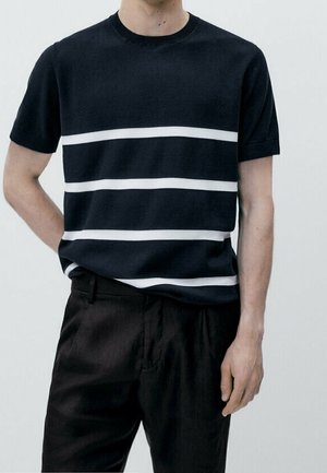 Mężczyzna w czarnej koszulce z krótkim rękawem z trzema białymi poziomymi paskami, w połączeniu z czarnymi spodniami, stojący na tle jednolitym.