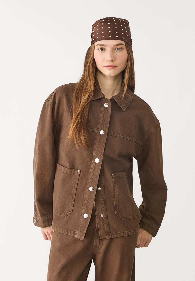 Brun denimjacka med en avslappnad passform, som har en tryckknappsstängning, två framfickor och en krage. Bärs med ett brunt hårband med prickar.