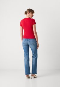 T-shirt rouge ajusté, à manches courtes, associé à un jean évasé bleu clair. Le modèle porte des mules à talons beiges, se tenant devant un fond uni.