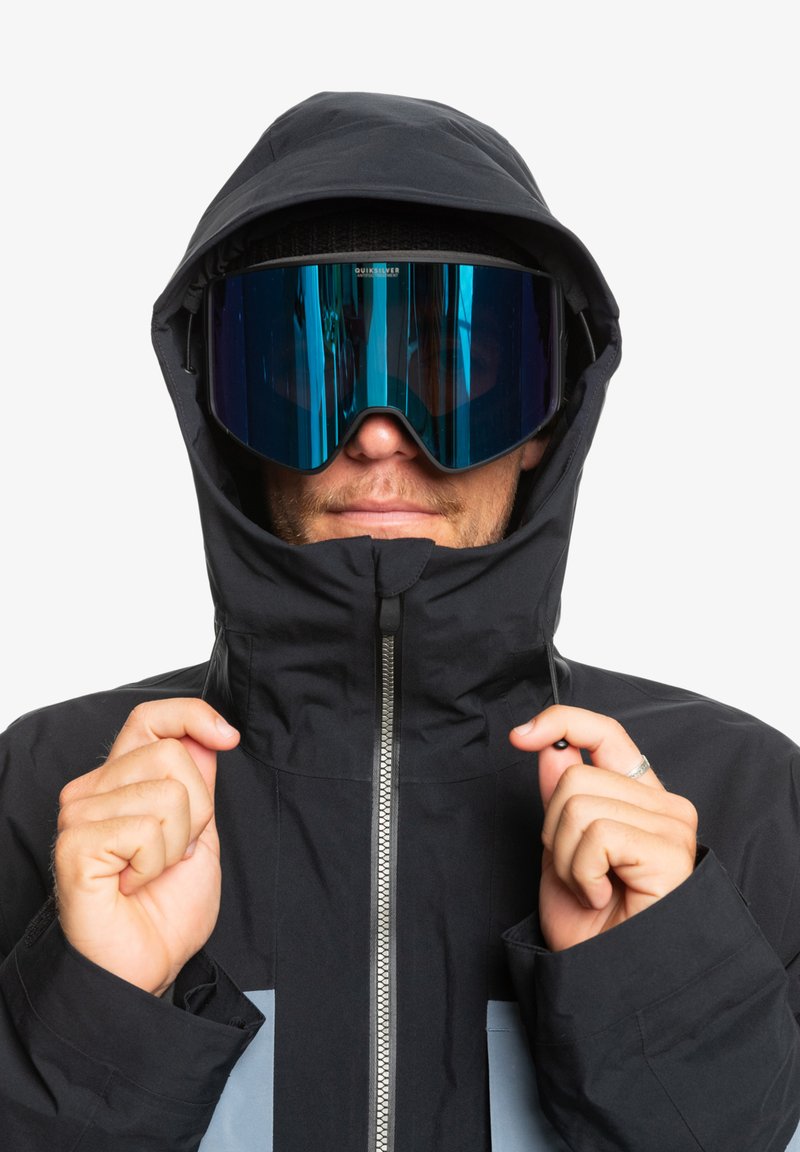 Czarna wodoodporna kurtka z wysokim kołnierzem i szarymi akcentami, w zestawie z dużymi gogle narciarskimi w niebieskim odcieniu. Posiada zamek błyskawiczny z przodu i kaptur.