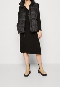 Mustapuffaliivi hupulla, jossa on vetoketjukiinnitys ja sivutaskut, käytetty pitkän mustan mekon päällä. Beige nilkkurit mustilla yksityiskohdilla.