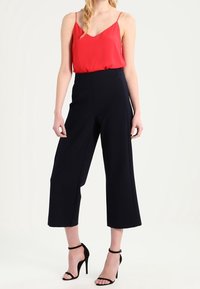 Donna che indossa un top rosso senza maniche, pantaloni neri a gamba larga e corta, e sandali neri con tacco alto e cinturino alla caviglia, in piedi contro uno sfondo bianco.