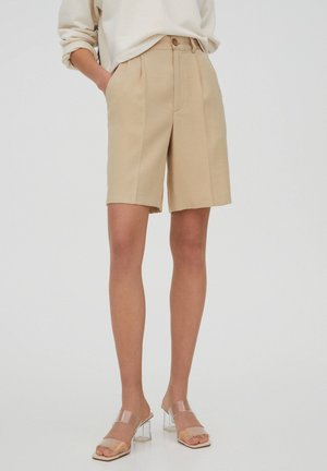 Femme portant un short plissé beige taille haute, un haut blanc à manches longues retroussées et des sandales transparentes à talons blocs, debout devant un fond blanc.