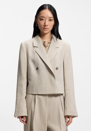 Donna con lunghi capelli neri che indossa un blazer corto doppiopetto beige e pantaloni a gamba larga a vita alta coordinati su sfondo bianco.