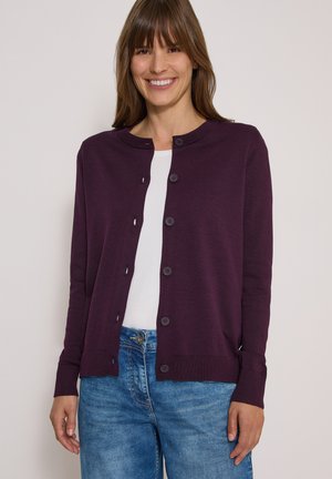 Femme souriante portant un cardigan violet boutonné par-dessus une chemise blanche et un jean bleu, debout devant un fond clair.