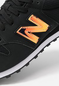 Svart atletisk sko med mesh- och syntetiskt överdrag, med en orange reflektiv logotyp, vit mellansula och svart gummisula med grepp.