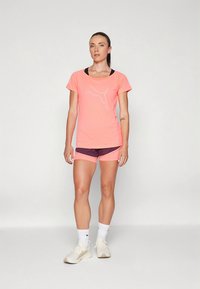 Roosa lühikeste varrukatega spordisärk, millel on valge logo. Sobitatud lilla lühikeste pükstega, valgete pahkluusokkidega ja beežide tossudega.