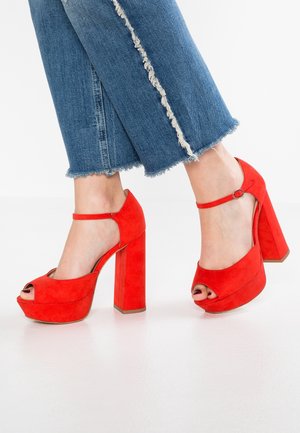 Red suede platform heels with a peep toe and ankle strap. They feature a chunky block heel and a smooth texture.