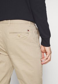 Beżowe spodnie z zapięciem na guzik z tyłu i małym logo w kolorze czerwonym, białym i niebieskim, noszone z czarną bluzką z długim rękawem, z luźno ułożoną prawą ręką.