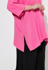 Maglietta fucsia over-size con maniche corte, caratterizzata da spacchi laterali e un orlo arrotondato, abbinata a pantaloni neri. Tessuto liscio.