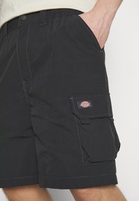 Black cargo shorts made of lightweight fabric, featuring front pockets, side cargo pocket with logo patch, and contrast stitching details.