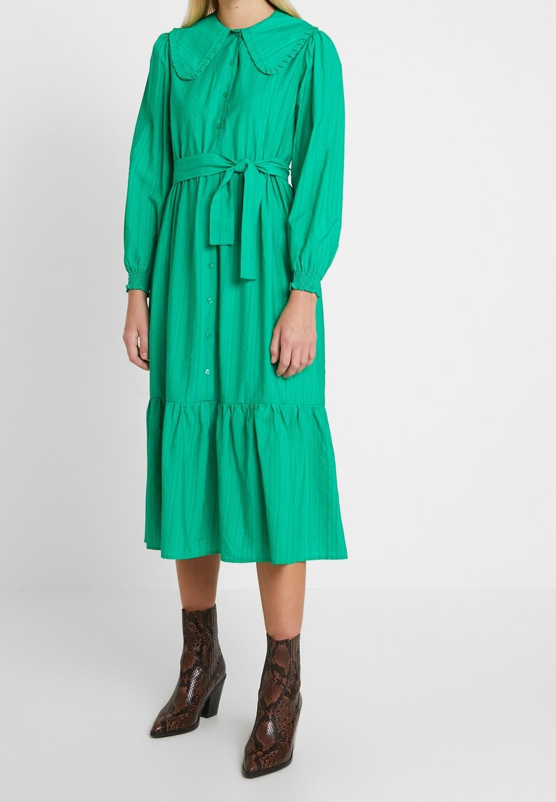 Femme portant une robe verte à manches longues avec des boutons sur le devant, un grand col, une ceinture à la taille et des bottines marron en peau de serpent.