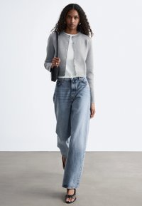 Kvinna som går framåt iförd en grå knappad cardigan, vit skjorta, vida jeans, svarta sandaler och bär en svart axelväska.