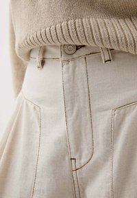 Beige gebreide trui over witte denim broeken met zichtbare zakken en contrasterende stiksels, die een textured breisel en een gladde stof toont.