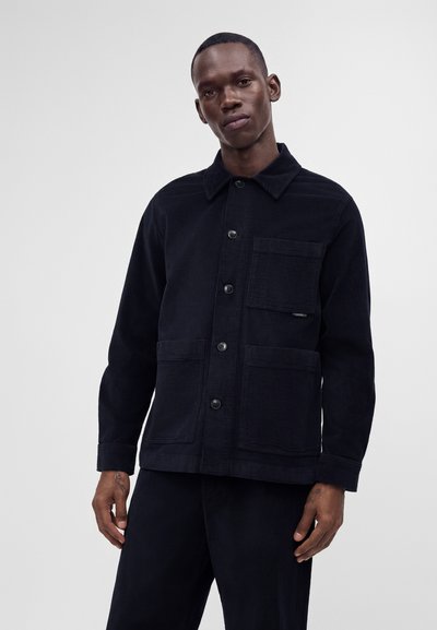 Homme portant une veste noire en velours côtelé à boutons avec trois poches avant, debout devant un fond clair uni.