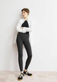 Zwarte legging met een gladde textuur en een hoge taille, gecombineerd met een nauwsluitend zwart topje en een wit overhemd met knopen. Draagt zwarte sneakers met witte veters.