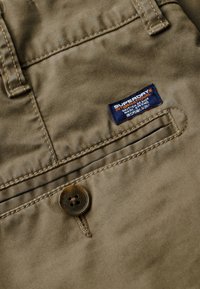 Beige bavlněné kalhoty s hladkou strukturou, s viditelným knoflíkem a modro-oranžovou značkovou nášivkou na zadní kapse.