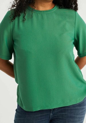 Personne aux cheveux bouclés portant un haut vert uni à manches courtes et un jean bleu, debout les mains derrière le dos.