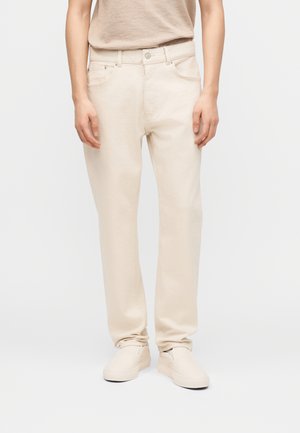 Isik, kes kannab helevalgeid lõtkvate jalgade teksasid, sobivaid slip-on jalanõusid ja helebeeži sisse toppitud pluusi, seisab ühevärvilise tausta ees.