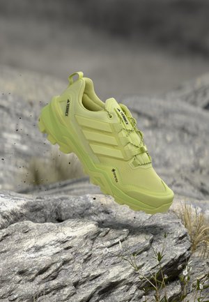 Chaussure de trail running Adidas Terrex Gore-Tex jaune flottant au-dessus d'un terrain rocheux avec de petites plantes et des particules de poussière autour.
