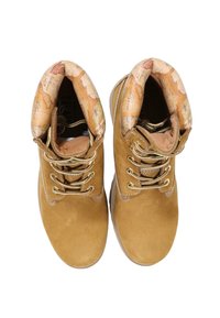 Botas de tobillo de ante marrón con ojales y cordones dorados. El interior presenta un diseño de mapa con patrones. La suela es lisa y de color claro.