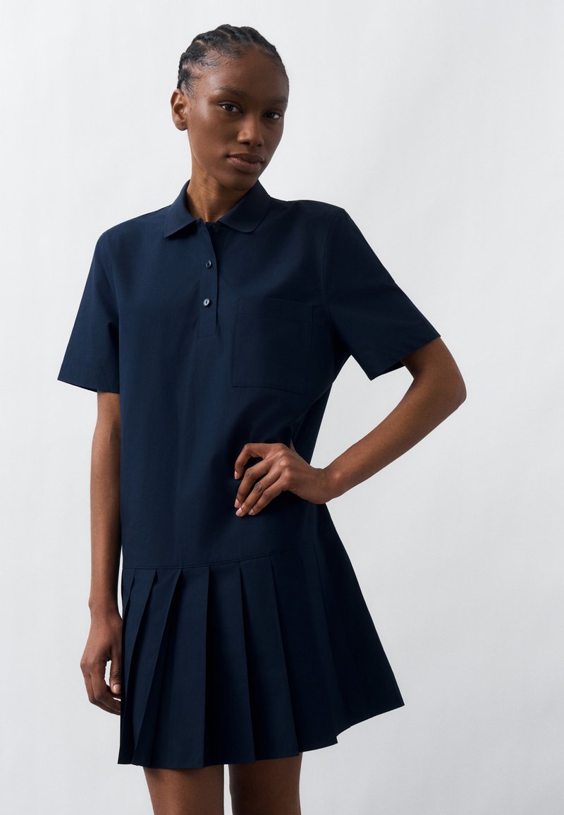 Γυναίκα με πλεξούδες στα μαλλιά, φορώντας φόρεμα πόλο κοντομάνικο σε ναυτικό μπλε με τσέπη στο στήθος και πλισέ φούστα, ποζάρει μπροστά σε απλό φόντο.