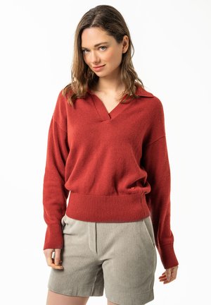 Woman wearing a rust-red V-neck sweater and light beige corduroy shorts, standing with a slight smile against a white background.