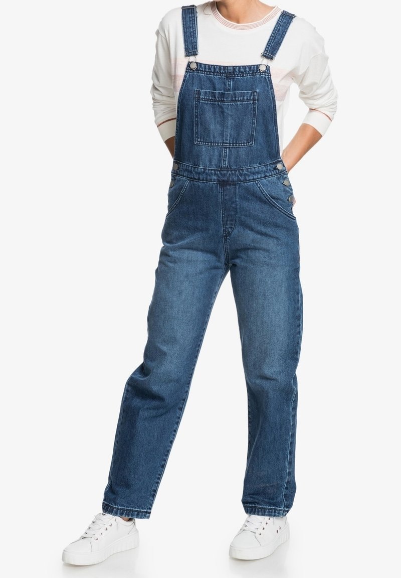 Denimbyxor i mörkblått med justerbara axelremmar, framficka och sidoknappar. Matchas med vita sneakers.