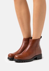 Braune Lederstiefeletten mit schwarzen Profilsohlen, getragen an nackten Beinen vor einem weißen Hintergrund.