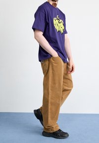 Lilla t-shirt med korte ærmer og gul grafisk print, kombineret med brune manchetter i kordfløjl og sorte sneakers. Enkelt, afslappet outfit.