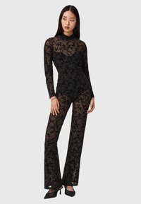 Black sheer jumpsuit with long sleeves, floral lace pattern, fitted bodice, and wide pants. Paired with black heeled shoes.