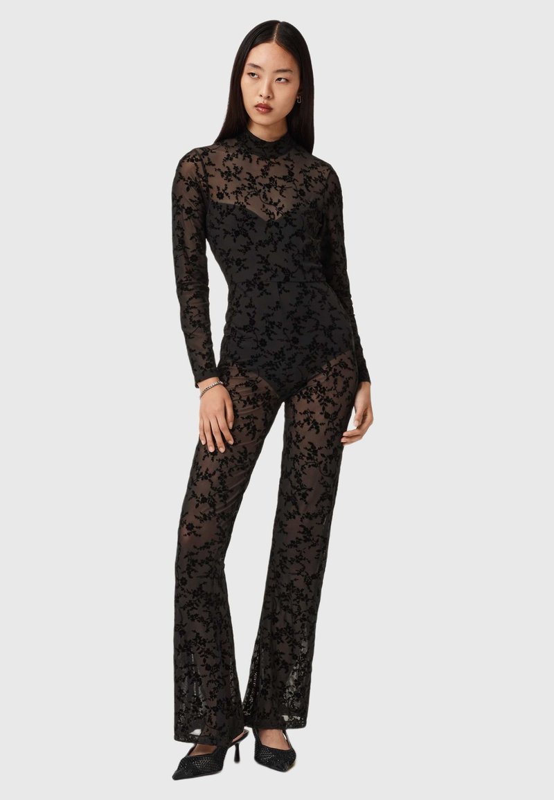 Black sheer jumpsuit with long sleeves, floral lace pattern, fitted bodice, and wide pants. Paired with black heeled shoes.