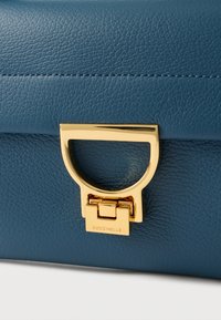 Blue leather handbag featuring a textured surface, gold hardware clasp, and a distinctive circular handle design with logo detail.