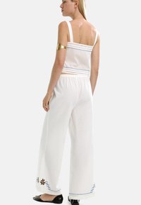 Kobieta ubrana w białą, bezrękawnikową bluzkę i szerokie spodnie w niebieskie i kwiatowe wzory, stojąca boso na białym tle.