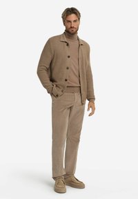 Tan stickad cardigan med knappar, lagd över en beige polokrage och beiga kordflorsbyxor. Skorna är ljusbruna slip-ons.