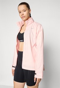 Light pink waterproof jacket with a high collar, zippered front, and reflective accents. Worn over a black sports bra and shorts.